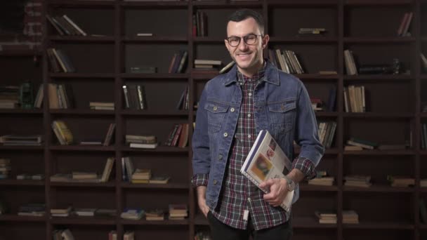 Barnman glad student håller bok framför bokhylla i biblioteket — Stockvideo