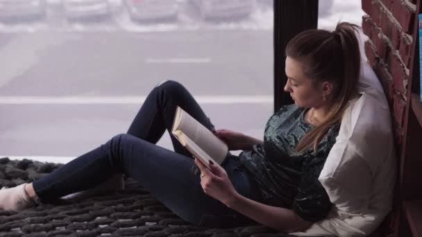 Young pensive woman reading book on bed by window overlooking city street — Stock Video