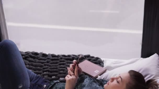 Mujer joven usando teléfono inteligente en la cama por ventana con vistas a la calle de la ciudad — Vídeos de Stock