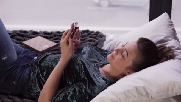 Jovem mulher usando smartphone na cama pela janela — Vídeo de Stock