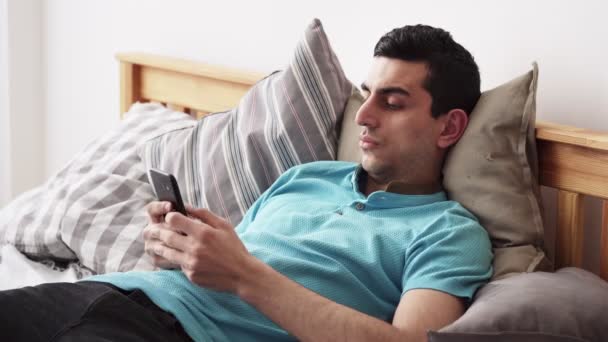 Молодой мужчина с Ближнего Востока, с помощью смартфона, лежит на кровати дома — стоковое видео