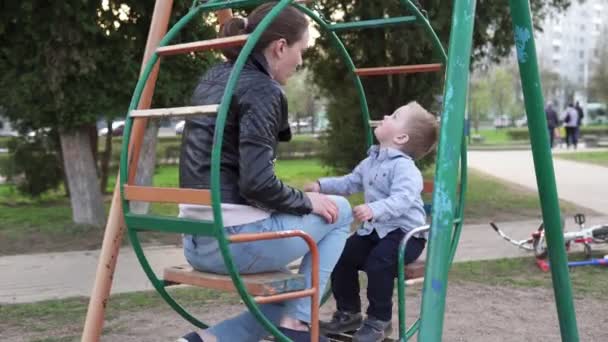 Mother and little curious son sitting together on a swing in park — Stock Video