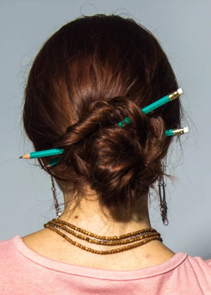 Pencils in head hair updo. Isolated back shot of brunet woman with two green pencils  in her hair. Girls unique hairstyle. Concept of style, student and beauty.