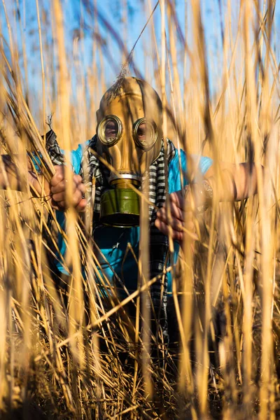 Man in blue t shirt and scarf Shemagh in gas mask sneaky sitting and spying in reeds field. Environmental protection of ecology. Concept of infection, apocalypse and pollution. Hope concept.