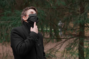 Siyah paltolu genç adam portresi. Siyah tıbbi maske takıyor. Parkta yürüyen klasik montlu bir adam. Koruyucu maskeli adam kopyalama alanı. Adam yukarı bakıyor, düşünüyor. Ormanda merak eden kişi. 