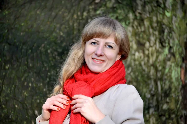 Die fröhliche junge Frau bindet sich einen roten Schal um. Porträt vor dem Hintergrund eines Baumes — Stockfoto