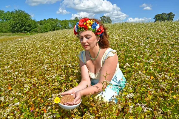 Die Frau mit dem Kranz auf dem Kopf hält eine Schale mit Buchweizen im Feld des blühenden Buchweizens — Stockfoto