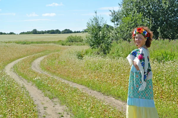 Kafasına bir çelenk ile neşeli kadın çiçek açması buğday alanında bir yolda gidiyor