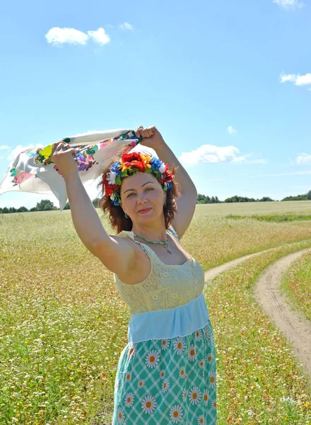 The mature woman with a wreath on the head holds a colorful scar in the raised hands against the background of the blossoming buckwheat field — Stock Photo, Image