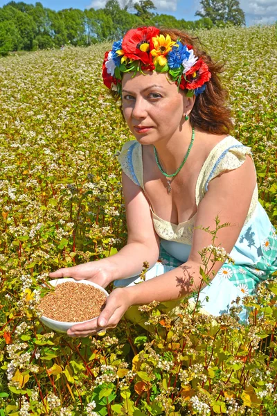 Žena v průměru let s věncem na hlavě drží misku s pohankou v oblasti kvetoucí pohanky — Stock fotografie