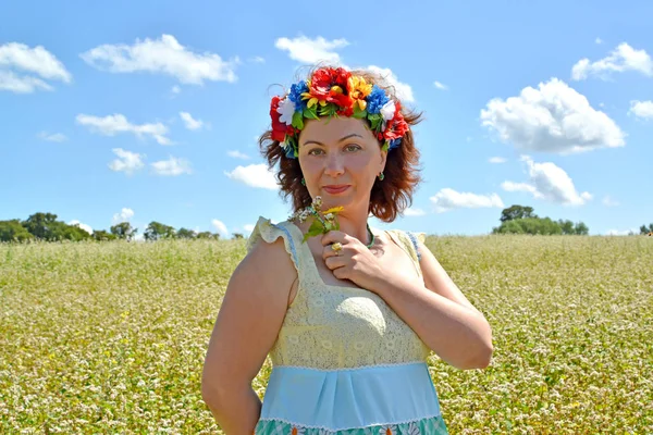 La femme d'années moyennes avec une couronne sur la tête tient des fleurs sur le fond du champ de sarrasin — Photo
