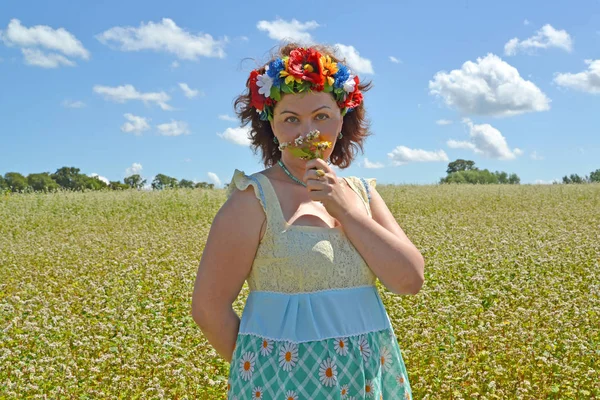 Die reife Frau mit einem Kranz auf dem Kopf riecht Blumen vor dem Hintergrund des Buchweizenfeldes — Stockfoto