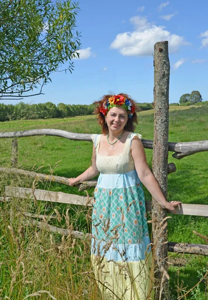 The cheerful woman of average years with a wreath on the head stands near a fence