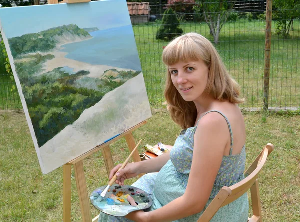 La joven artista embarazada pinta un cuadro, sentada en un caballete — Foto de Stock