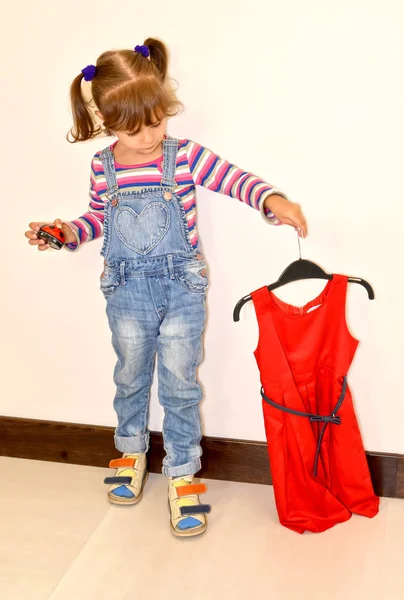 The little girl holds a beautiful red dress on a hanger — Stock Photo, Image