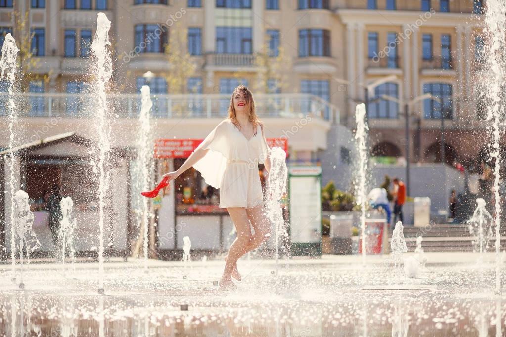 Young girl in a light dress is having fun in a fountain on the b