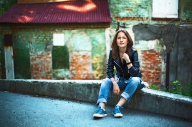 Bir kız bir deri ceket, kot pantolon ve spor ayakkabı içinde; R oturan