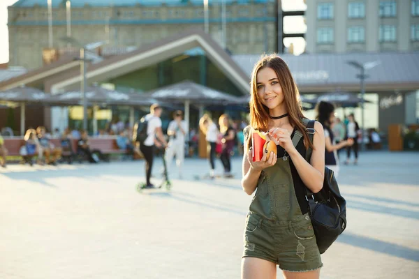 A young girl with a cheeseburger and fries in her hand walks aro — Stock Photo, Image