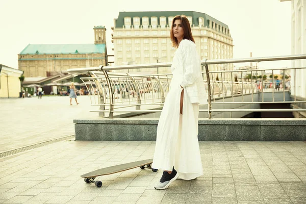 Girl in a white costume stands with a longboard on the square of — Stock Photo, Image