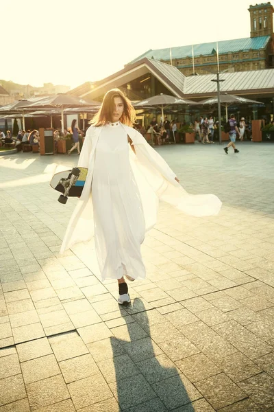 Fille dans une robe blanche et avec un longboard sur la place ensoleillée o — Photo