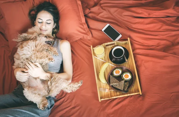 Ung kvinna ligger i sängen med hund och att ha en hälsosam frukost. — Stockfoto
