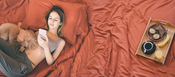 Ung kvinna är liggande med hund i sängen och att ha en hälsosam frukost. — Stockfoto