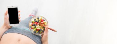 Genç kadın bir meyve salatası yemek yiyor ve fitness app üstünde onun smartphone bir egzersiz yaptıktan sonra kullanarak..