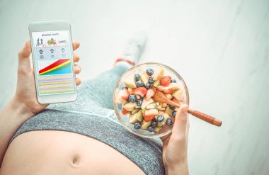 Genç kadın bir meyve salatası yemek yiyor ve fitness app üstünde onun smartphone bir egzersiz yaptıktan sonra kullanarak..