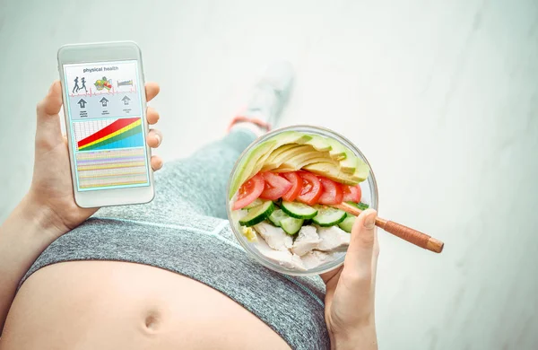 Junge Frau isst Salat und bedient sich nach dem Training einer Fitness-App auf ihrem Smartphone. — Stockfoto