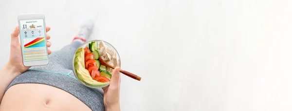 Mujer joven está comiendo una ensalada y utilizando una aplicación de fitness en su teléfono inteligente después de un entrenamiento . — Foto de Stock