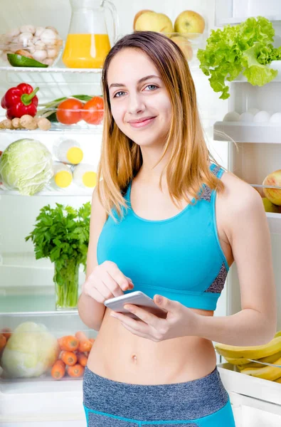 Mujer joven sonriente usando una aplicación para teléfonos inteligentes y manteniéndose cerca de la nevera llena de comida saludable . —  Fotos de Stock