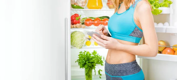 Mujer joven usando una aplicación para teléfonos inteligentes y manteniéndose cerca de la nevera llena de comida saludable . —  Fotos de Stock