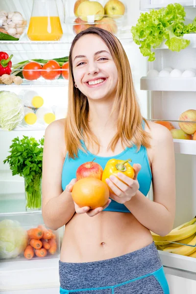 Mujer joven sonriente con frutas y verduras cerca de la nevera llena de comida saludable . —  Fotos de Stock
