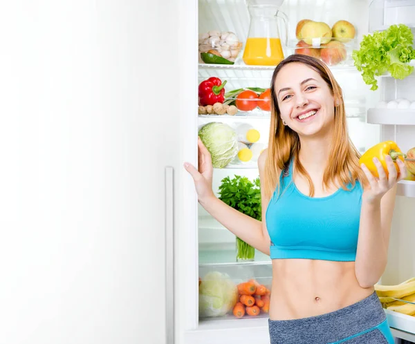 Smiling young woman staing with fruits and vegetables near the fridge full of healthy food. — Stock Photo, Image