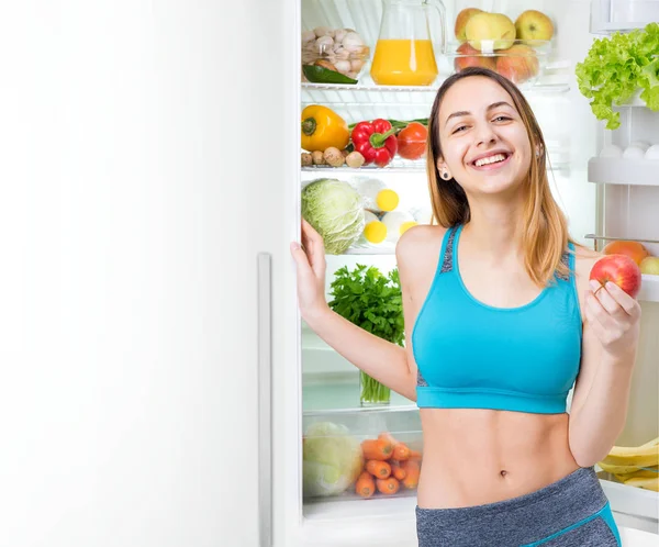 Sorrindo jovem mulher comendo uma maçã e ficar perto da geladeira cheia de alimentos saudáveis . — Fotografia de Stock