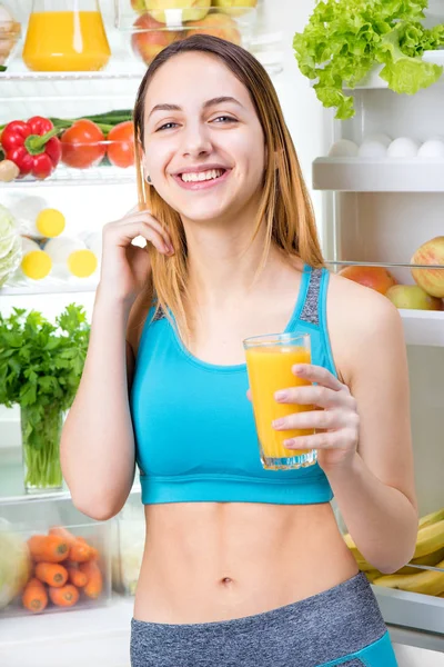 Jovem mulher bebendo suco e ficar perto da geladeira cheia de alimentos saudáveis . — Fotografia de Stock