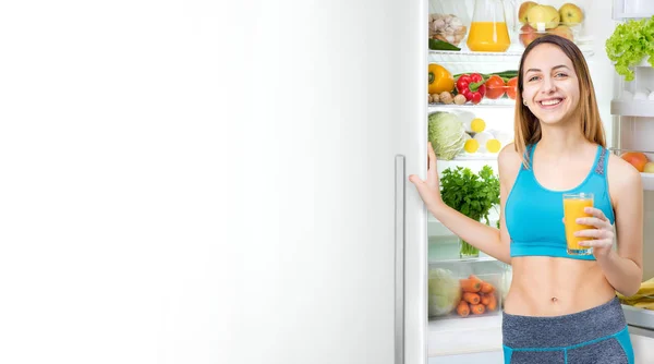 Young woman drinking juice and staying near the fridge full of health food. — Stock Photo, Image