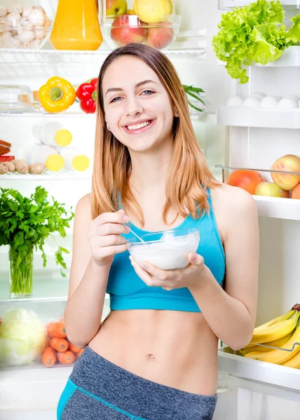 Young woman eating a yogurt and staying near the fridge full of health food. — Stock Photo, Image