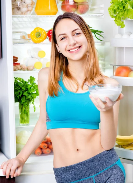 Young woman eating a yogurt and staying near the fridge full of health food. — Stock Photo, Image
