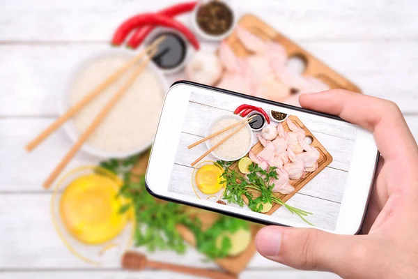 Manos tomando fotos de alitas de pollo frescas con especias y tazón con arroz con smartphone . — Foto de Stock