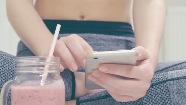 Giovane donna sta bevendo un frullato e utilizzando un'applicazione fitness sul suo smartphone dopo un allenamento . — Video Stock