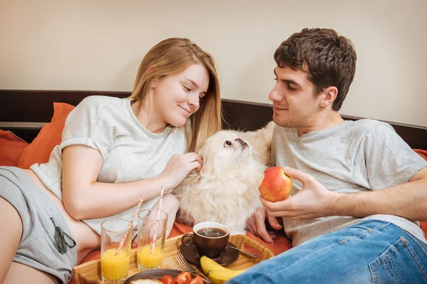 对年轻的夫妇躺在床上与狗和吃健康的早餐. — 图库照片