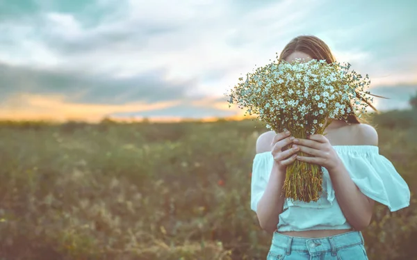 Vacker flicka står i ett fält som håller en bukett av vita blommor i sina händer. — Stockfoto