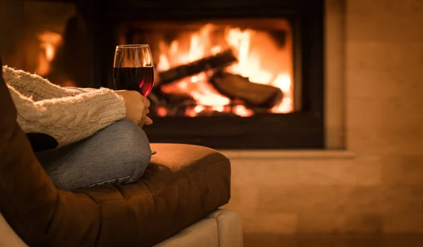 Jovem mulher sentada em casa junto à lareira e bebendo um vinho tinto . — Fotografia de Stock