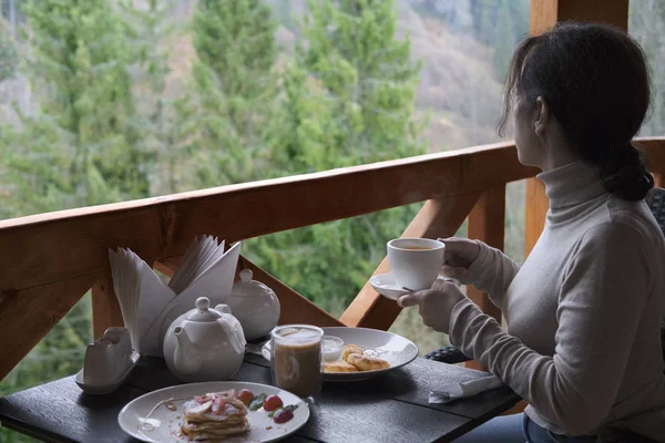 这个年轻的女人坐在阳台上吃早饭 背景美丽的山脉 — 图库照片