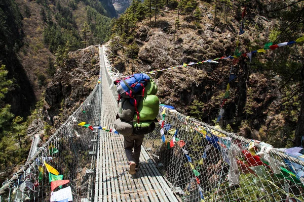 Nepal Porter carrying heavy load on his back going along the bridge