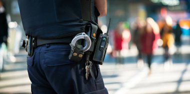 Polis memuru insanları izliyor. Arka planda kelepçeli bir polis ve bir grup bulanık insan var..