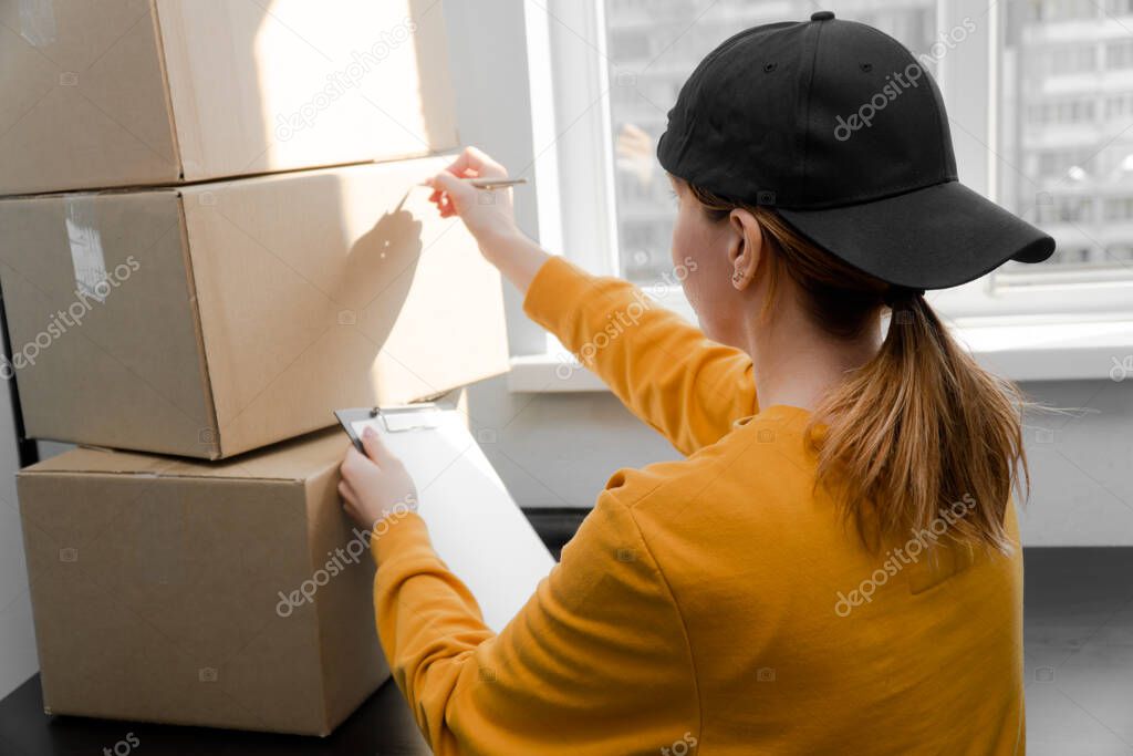 Girl storekeeper in a black cap signs a cardboard paper box with the goods. The courier takes the order from the customers house. side view on a light background