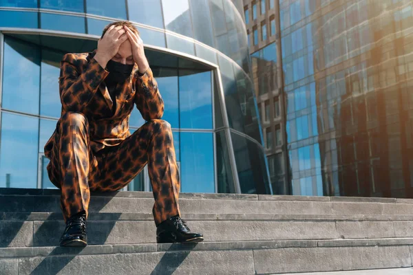 Portrait of an angry european man in a mask on the background of a business center. average plan businessman is sitting on the steps in a business suit.