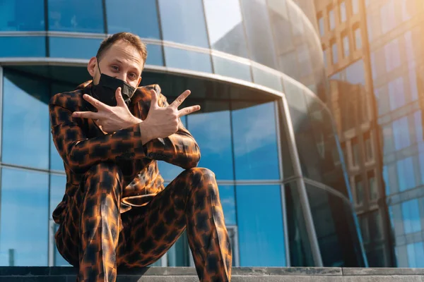 Portrait of a cheerful European man in a medical mask on the background of a business center. middle plan businessman indulges on the steps in a business suit.